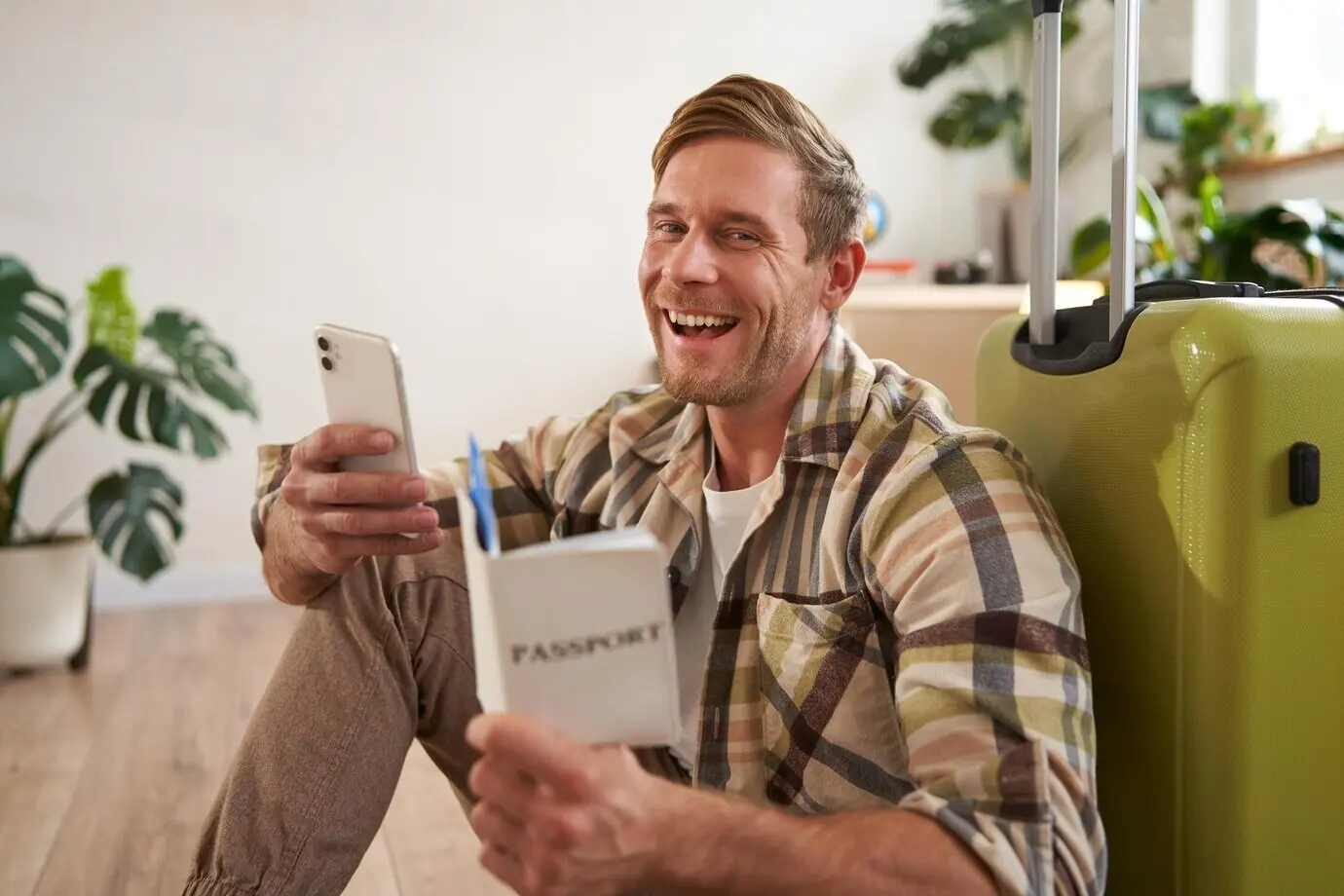 A portrait of a smiling, handsome male tourist holding a passport and flight tickets, sitting with a suitcase.