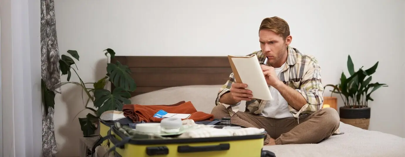 Portrait of an angry man packing a suitcase, looking frustrated at a notebook with a checklist of items for a holiday, preparing to go on vacation.